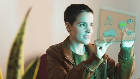 Sanft lächelnde Person mit Nasenpiercing hält beide Hände auf vor sich befindlichem grünblau leuchtendem Hologramm mit verschiedenen Grafiken: Baum, Uhr, Auto, Blitz und Figur auf Tretroller