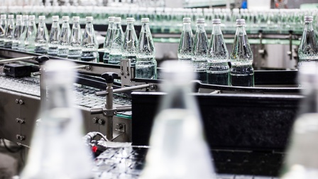 Mehrere transparente Glasflaschen mit transparenter Flüssigkeit abgefüllt auf Rollband im Fokus, im Vorder- und im Hintergrund verschwommen weitere Flaschen