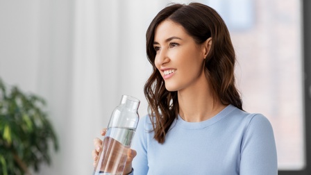 Frontale Aufnahme einer Person mit dunklen, offenen Haaren und hellblauem Pullover, die aus einem Glasbehälter Wasser trinkt. Im Hintergrund ein Fenster, eine Zimmerpflanze und weiße Vorhänge