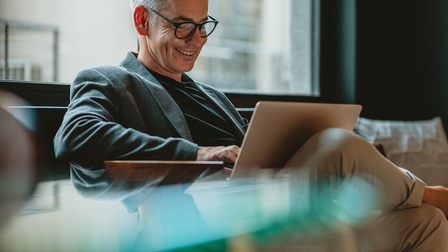 Person mit kurzen grauen Haaren und Brille sitzt mit einem Laptop auf dem Schoß und blickt freudig darauf