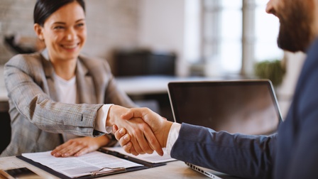 Zwei Personen sitzen sich an einem Tisch gegenüber und schütteln sich die Hände. Auf dem Tisch liegen ein Clipboard, ein aufgeklappter Laptop, ein Smartphone