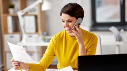 Person mit kurzen dunklen Haaren sitzt an einem Schreibtisch mit Laptop, telefoniert mit einem Smartphone und blickt dabei auf Unterlagen, die in der Hand gehalten werden, im Hintergrund zeigt sich eine Büroräumlichkeit