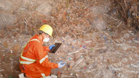 Person mit orange-gelber Schutzbekleidung kniet in einem Raum mit leeren Plastikflaschen und hält dabei ein Tablet sowie eine Pet-Flasche in der Hand