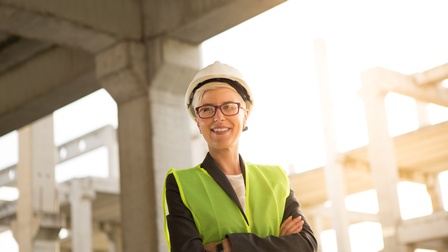 Person mit kurzen Haaren, Businesskleidung und Brille steht mit gelber Warnweste und verschränkten Armen und weißem Schutzhelm auf einer Baustelle