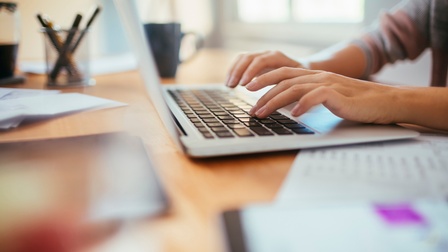 Eine Person tippt auf der Tastatur eines Laptops am Schreibtisch. Daneben liegen Zettel, Stifte und eine Kaffeetasse.