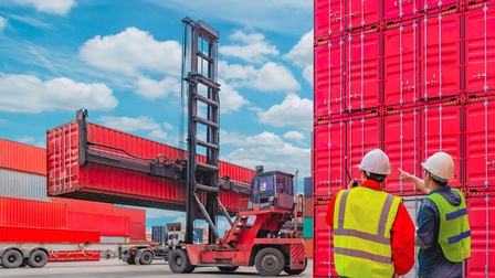 Zwei Personen mit gelben Warnwesten und weißem Schutzhelm blicken auf einen Containerstapler, der Container umsetzt, daneben stehen weitere rote Container gestapelt