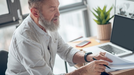 Bärtige Person sitzt an Schreibtisch vor aufgeklapptem Notebook und sichtet Stapel Unterlagen