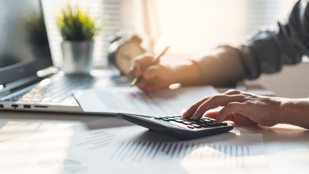 Eine Person sitz am Schreibtisch und kalkuliert mit einem Taschenrechner 