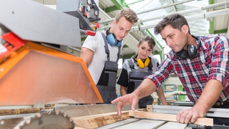 Person steht bei einer Kreissäge in einem Holzverarbeitungsbetrieb und zeigt zwei weiteren jugendlichen Personen die Verarbeitung von einem Holzstück