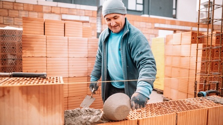 Person mit grauer Haube, blauem Pullover, Jacke und Handschuhen baut eine Ziegelwand