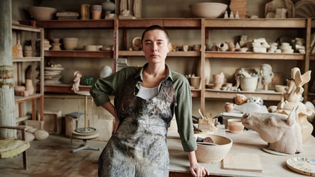 Person n Schürze steht mit einer Hand an Tisch gelehnt in Töpferwerkstatt, im Hintergrund Regale voller Töpferwaren