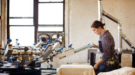 Person in Schürze mit Farbflecken bedient Siebdruckmaschine, im Hintergrund Metallrohre und Fenster