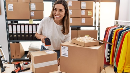Lächelnde Person steht vor Tisch mit Kartonagen und scannt Code auf einer der Kartonagen, im Hintergrund Regal mit weiteren Paketen und behängte Kleiderstande