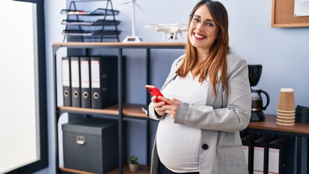 Lächelnde, schwangere Person mit Brillen steht in Büroraum und hält rotes Smartphone in Händen. Im Hintergrund verschwommen Regal mit Aktenordnern Schachteln und einer Kaffemaschine.