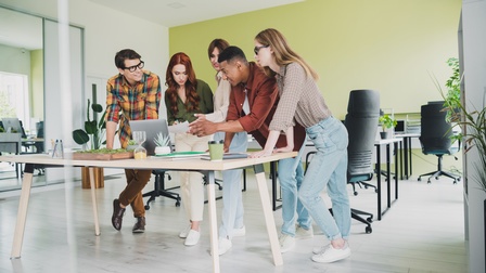 In einem Büro stehen um einen Tisch fünf Personen. Auf dem Tisch ist ein Laptop, auf den zwei Personen deuten. Eine dritte Person notiert etwas auf einem Zettel. Im Hintergrund sind Schreibtische und Sessel