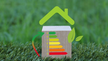 Ein kleiner Holzwürfel steht auf grünem Gras. Auf dem Würfel ist die Grafik eines Balkendiagramms, die Balken werden von oben nach unten länger und stellen farblich Energieklassen dar. Darüber symbolisch ein grünes Dach, links ein Pfeil, rechts ein Blatt.