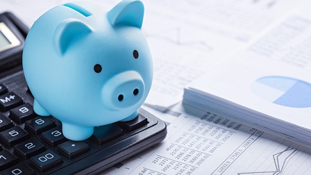 Blaues Sparschwein steht auf einem Taschenrechner neben verschiedenen Unterlagen mit Zahlen, Diagrammen und Kurven