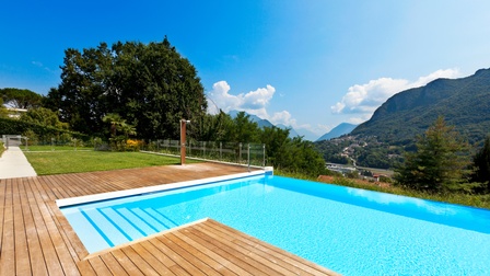 Ein großes Becken mit klarem Wasser, in das Stiegen an einer Stelle führen, wird von Holzbrettern umgeben. Links von den Brettern ist eine grüne Rasenfläche. Im Hintergrund sind grüne Berge und Häuser