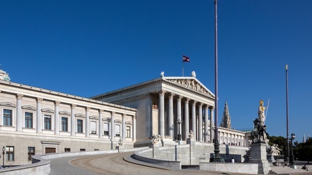 Aufnahme der Vorderseite des Parlaments in Wien. Vor dem Parlament ist ein Brunnen. Der Himmel ist blau. Im Hintergrund sind grüne Bäume sowie Turmspitzen