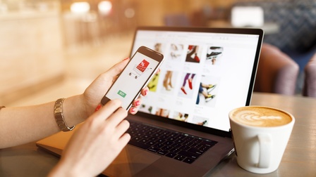Person sitzt vor einem Laptop und kauft über ein Smartphone ein