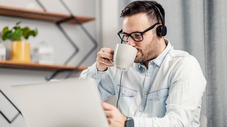 Person mit Headset und Brillen nimmt Schluck aus weißer Tasse und sitzt vor aufgeklapptem Notebook, im Hintergrund verschwommen graue Gardine und Regale mit Pflanze in gelben Topf