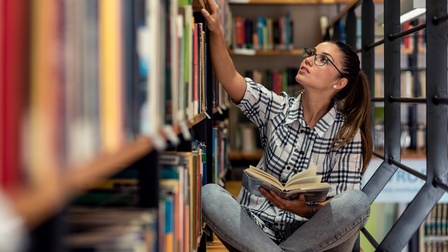 Person mit Brillen sitzt am Boden einer Bibliothek, hält in einer Hand aufgeschlagenes Buch und langt mit anderer Hand in Bücherregal darauf blickend