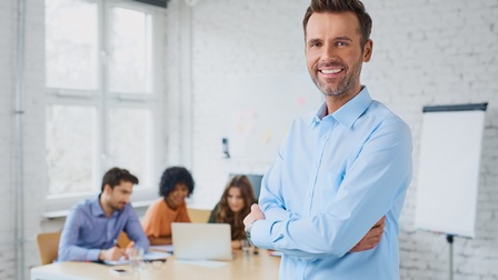 Porträt einer Person mit hellblauem Hemd, kurzen Haaren, Bart und verschränkten Armen, die in einem großen, hellen Büroraum steht. Im Hintergrund sitzen drei Personen an einem Tisch. Vor ihnen steht ein Laptop, auf den alle drei blicken
