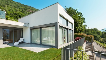Weißes quadratisches Haus mit großen Fensterflächen im Sonnenlicht unter blauem Himmel, im Hintergrund grün bewachsener Hügel, Sträucher und Bäume. Vor dem Haus Steinterrasse mit weißer Liege in Wiese verlaufend, mit Geländer eingefasste Treppe.