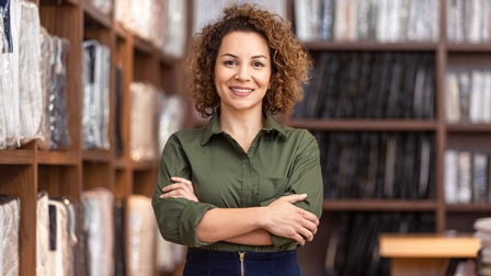 Person mit lockigen, schulterlangen Haaren und  grüner Bluse steht mit verschränkten Armen freudig vor Regalen mit Bettwaren