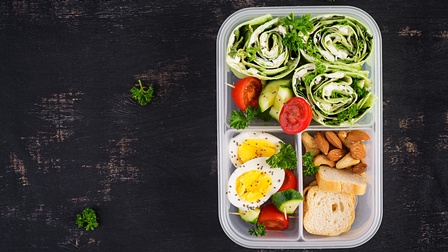 Top-Shot einer Plastikbox in der rechten Hälfte mit drei Fächern. In jedem Fach ist unterschiedliches Essen, zum Beispiel Gurken, Tomaten, Nüsse, Zwieback, Wraps mit Gemüse