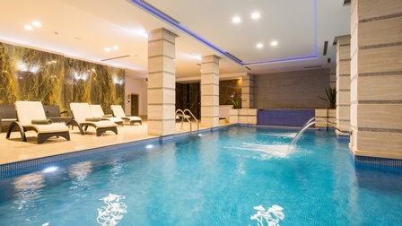 Ein Hallenbad, ausgelegt mit blauen, kleinen Kacheln in Form eines Rechtecks. Links neben dem Wasser stehen auf einer Fläche mehrere Liegen. Auf manchen Liegen ist ein zusammengerolltes Handtuch.