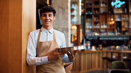 Porträt einer Person in einer hellbraunen Schürze, die sich in einem Lokal an einer Säule anlehnt und ein Tablett hält. Im Hintergrund ist eine Bar mit einem Tresen