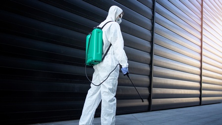 Person in Rückenansicht in weißem Schutzanzug mit Schutzbrille, Atemschutz und Handschuhen trägt türkisen Container verbunden mit Sprühstab am Rücken, vor Lamellenwand stehend