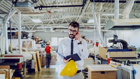 Frontale Aufnahme einer Person mit Hemd, Brille und Krawatte, die in einer Werkstatt mit Druckerpressen steht und telefoniert. Die Person hält ein aufgeklapptes Notizbuch. Im Hintergrund sind weitere Personen sowie verschiedene Drucke