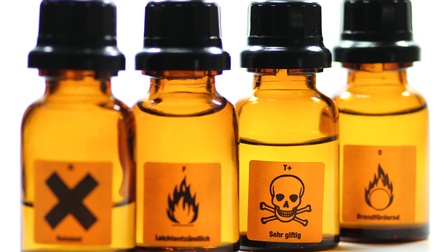 Braune kleine Glasfläschchen mit schwarzen Verschlüssen und Etiketten mit schwarzem X, Flammen und Totenkopf, Fokus auf Letzterem vor weißem Hintergrund