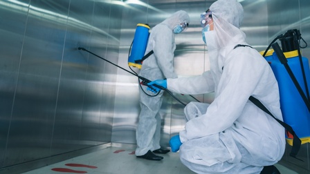 Zwei Personen befinden sich mit Schutzkleidung und Masken in einem geschlossenen Aufzug und desinfizieren diesen.