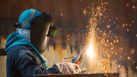 Person in Schutzbekleidung mit Schutzbrille und -Handschuhen führt Schweißerarbeiten durch und Funken erleuchten den Raum