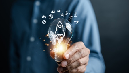 Eine Person hält eine Glühbirne in die Kamera. In der Glühbirne sind die Umrisse einer Rakete. Am unteren Ende ist die Glühbirne beleuchtet. Über der Glühbirne sind grafische Symbole wie eine Zielscheibe, Zahnräder, eine Checkliste oder Puzzleteile