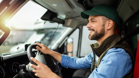 Seitliche Aufnahme einer hinter einem Lenkrad eines Autos sitzender Person, die ihre Hände auf dem Lenkrad hat und aus der Windschutzscheibe blickt. Die Person trägt eine dunkelgrüne Kappe