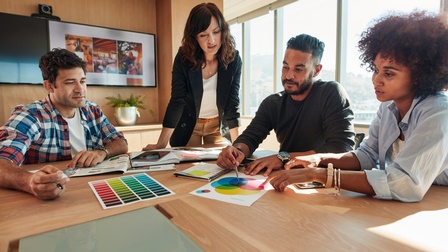 Personen sitzen und stehen an einem Tisch in einem hellen Büro und betrachten Farbkarten