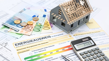Dokumente mit Schriftzug Energieausweis für Wohngebäude verstreut, daraufliegend Taschenrechner, Euro-Geldscheine und -Münzen sowie kleines Modell eines Hauses