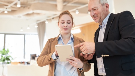 Zwei Personen stehen nebeneinander und blicken auf ein Tablet, im Hintergrund ist ein modernes und hell beleuchtetes Büro