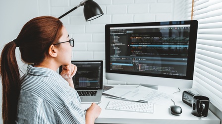 Person mit Brillen stützt Hand an Kinn an Schreibtisch sitzend und blickt auf Bildschirm, auf dem Quellcodes zu sehen sind