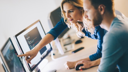 Zwei Personen blicken auf Computermonitor, eine Person bedient Maus, die andere Person deutet mit Finger auf Monitor