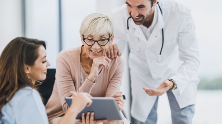 Medizinisches Personal in weißer Arbeitskleidung bespricht mit einer Person mittleren Alters und hellen, kurzen Haaren mit Brille etwas und blicken dabei auf ein Tablet