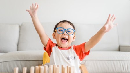 Kind mit dunklen kurzen Haaren und Brille sitzt am Boden vor einem hellen Sofa und streckt freudig die Hände in die Höhe, davor stehen Holzklötze auf einem Couch-Tisch