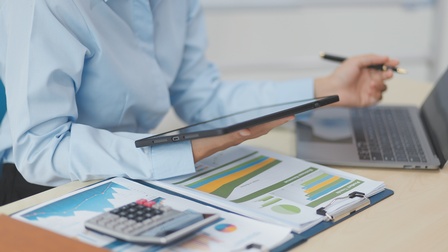 Eine Person in einer hellblauen Bluse sitzt an einem Schreibtisch. Mit der rechten Hand hält sie ein Tablet. Vor ihr auf dem Schreibtisch liegen Zettel mit verschiedenen Diagrammen. Auf ihnen liegt ein Taschenrechner