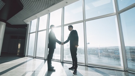 Zwei Personen in Anzügen stehen  vor einer Glasfront und schütteln sich die Hände. Durch die Glasscheiben scheint die tiefstehende Sonne