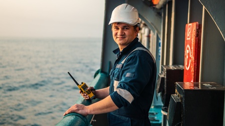 Person in Arbeitsoverall mit weißem Helm steht an Reling eines Bootes und hält Walkie Talkie in der Hand, im Hintergrund verschwommen Meer