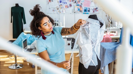 Person mit lockigem dunklen Haar steht bei einer Schneiderpuppe mit drapierten Stoffmuster und misst mit einem Maßband